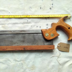 Op de afbeelding: Een vintage handzaag met een houten handvat en zaagblad. Het zaagblad is rechthoekig en heeft een donkere, verouderde uitstraling. De handgreep is gemaakt van lichtgekleurd hout en is bevestigd met messing klinknagels. Een gele rolmaat staat op de achtergrond.