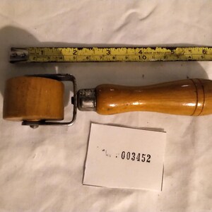 May include: A wooden seam roller tool with a cylindrical wheel and a smooth handle, featuring a metal bracket. A yellow measuring tape positioned above shows the tool is approximately 18 cm long. A small white tag displays the number 003452.