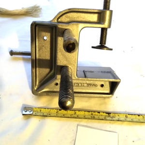 May include: A gray metal C-clamp with a threaded screw mechanism and a T-handle. A yellow measuring tape is positioned below the clamp, showing a width of approximately 6 inches. A small white tag with the number 003430 is visible.