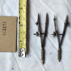 May include: Two silver-toned metal drafting compasses with knurled handles and adjustment screws, positioned next to a yellow measuring tape showing approximately 3.25 inches in length and a rectangular brown tag with the number 003179.