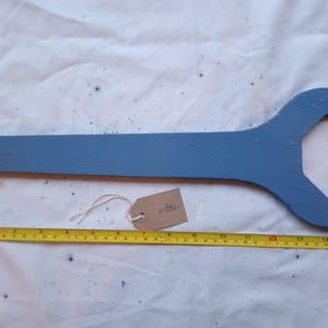 May include: A large, blue wooden wrench with a hole at the end of the handle. A yellow tape measure is placed next to the wrench, showing a length of approximately 38 cm. A small tag is attached to the wrench with a string.