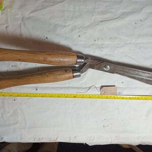 May include: Large, vintage hedge shears with wooden handles and a metal blade. The shears are open, and a yellow measuring tape is placed next to them. The handles are approximately 30 cm long.