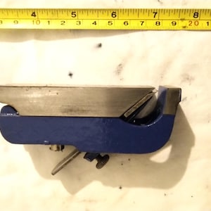 May include: A blue and silver metal hand plane with a wooden handle. The plane is approximately 15 cm long, with a blade and adjustment knob. A yellow measuring tape is in the background.