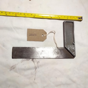 May include: A metal try square and a yellow tape measure are displayed on a white surface. The try square is a right-angled tool, and the tape measure shows inch measurements. A small tag with the number "902355" is attached.