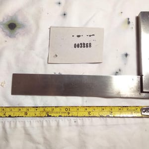 May include: A silver-colored metal machinist square positioned above a yellow measuring tape that shows a length of approximately 8 inches. A small white rectangular tag is placed between the arms of the square with the black printed text 003268.