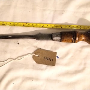 以下が含まれることがあります： 木製のハンドルと金属製の刃が付いた、ヴィンテージの木工用ノミ。 黄色の巻尺がノミの隣にあります。 番号002311の小さなタグがノミに付いています。