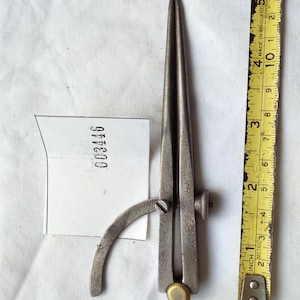 May include: A vintage metal drafting compass with a weathered grey finish, featuring a small brass knob at the pivot point and an adjustable curved arm. A white paper tag with the printed number 003446 is placed next to a yellow measuring tape.