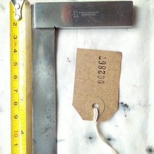 May include: A vintage metal try square with a blade measuring approximately 10 cm, next to a yellow measuring tape. The try square has the text "E. PRESTON & SONS, BERNS, ENG." and a brown tag with the number "002867".