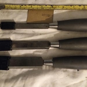 May include: Three metal chisels with grey handles and black protective caps on the blades, positioned below a yellow measuring tape that shows a length of approximately 10 inches. The tools feature metallic shafts and smooth, rounded grey grips.