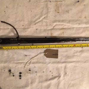 May include: A dark metal tool with a curved arm and a long, pointed blade, possibly for measuring or marking. A yellow tape measure is placed alongside the tool, with a tag attached. The tool appears to be approximately 46 cm long.