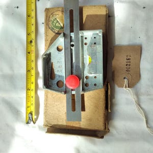 May include: A metal and plastic tool with a red knob, a level, and a ruler, resting on a cardboard box. A yellow tape measure is next to the tool. The ruler is approximately 20 cm long.