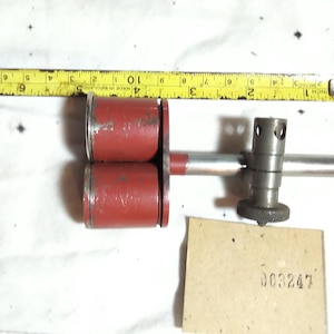 Puede incluir: Una herramienta de metal con dos componentes cilíndricos rojos unidos a una varilla horizontal con una perilla de ajuste plateada. Una cinta métrica amarilla arriba muestra una longitud de 11,5 cm. Una pequeña tarjeta beige muestra 003247.