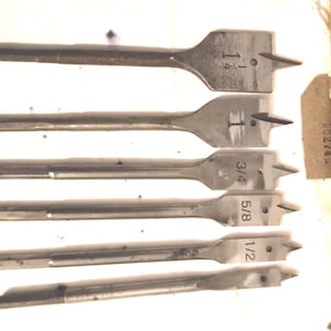 May include: A set of six metal spade drill bits of varying sizes, including 1/2, 5/8, 3/4, and 1 1/4 inch. Each bit has a flat, paddle-shaped head with a central point and cutting edges. A yellow measuring tape is next to the bits.