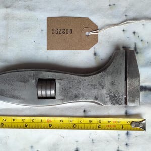 May include: A vintage adjustable wrench with a silver finish, lying next to a yellow tape measure. The wrench has a curved handle and an adjustable jaw. A brown tag with the number "362798" is attached to the wrench.