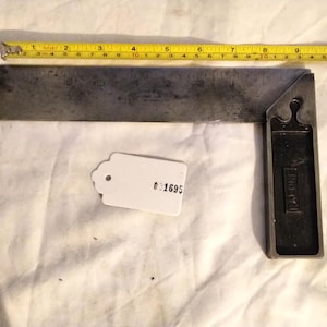 May include: A vintage metal try square with a 10-inch blade, a black handle, and a yellow measuring tape. A white tag with the number 01695 is also present. The tools are on a white fabric surface.