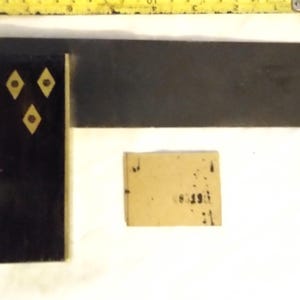 May include: A dark wood and metal try square with three brass diamond-shaped inlays on the handle, positioned below a yellow measuring tape showing approximately 23 cm, accompanied by a small rectangular tan paper tag with black text.