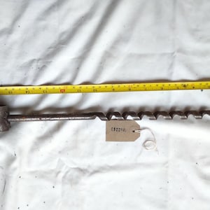 May include: An antique auger drill bit with a cylindrical handle and a spiral cutting edge. A yellow tape measure shows the drill bit is approximately 51 cm long. A tag with the number "C82203" is attached.