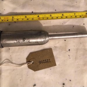 Puede incluir: Una pistola de engrase de metal vintage con una boquilla larga, un extremo roscado y una etiqueta con el número "002521". Una cinta métrica amarilla está al lado de la pistola de engrase.