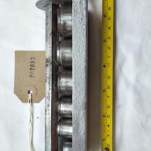 Könnte beinhalten: Ein vintage, silberfarbenes Metallwerkzeug mit sechs zylindrischen Kammern. Ein gelbes Maßband zeigt, dass das Werkzeug etwa 20 cm lang ist. Ein kleines Etikett mit der Nummer "002089" ist angebracht.