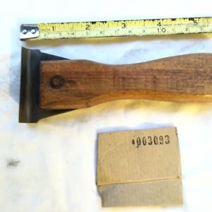 Könnte beinhalten: Ein Farbschaber mit Holzgriff und Metallklinge, platziert unter einem gelben Maßband, das eine Länge von etwa 15 cm anzeigt. Ein kleines beiges Pappquadrat mit der schwarz aufgedruckten Nummer 4003093 ist ebenfalls enthalten.