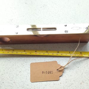 May include: A vintage wooden level with brass detailing and a yellow measuring tape. The level is approximately 25 cm long. A small brown tag with the number "001461" is attached.