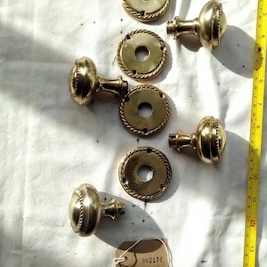 以下が含まれることがあります： アンティーク真鍮製のドアノブとバックプレートのコレクション。ノブは丸みを帯びた形状で、ロープのような装飾が施されています。バックプレートも同様のロープデザインの円形です。メジャーはセンチメートルを示しています。