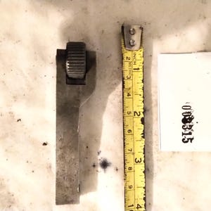 Pode incluir: Uma ferramenta de recartilhar em metal cinzento com uma roda cilíndrica texturada no topo, posicionada ao lado de uma fita métrica amarela com cerca de 10 cm de comprimento e uma etiqueta de papel branco com o número impresso 000315.