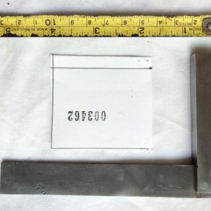 May include: A steel machinist square positioned next to a white square paper card featuring the black printed number 003462. A yellow measuring tape is placed above the items, showing a length of approximately 15 cm for the square tool.