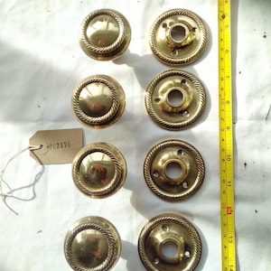 Peut inclure: Huit boutons de porte et plaques arrière en laiton vintage. Les boutons ont une forme arrondie avec une bordure en forme de corde. Les plaques arrière ont un design circulaire avec trois trous de vis. Un mètre ruban est à côté des boutons. Une étiquette avec le numéro "002170" est attachée.