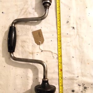 May include: A vintage hand drill with a black handle and a metal frame. The drill is approximately 38 cm long, with a brown handle at the top. A yellow measuring tape is next to the drill. A small tag is attached to the drill.