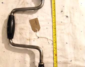 Op de afbeelding: Een vintage handboor met een zwarte handgreep en een metalen frame. De boor is ongeveer 38 cm lang, met een bruine handgreep aan de bovenkant. Een gele meetlint ligt naast de boor. Een klein label is aan de boor bevestigd.
