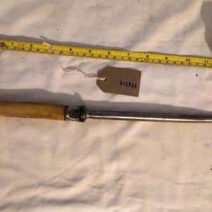 May include: A steel knife sharpener with a light-coloured handle and a silver-coloured metal bolster. A yellow tape measure is next to the sharpener. A small tag with the number "801934" is attached to the tape measure.