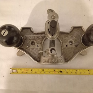 May include: A vintage Stanley No. 71 router plane, made in England, with a silver metal body and two dark wooden handles. A yellow measuring tape is placed below the plane, showing measurements in inches and centimeters.