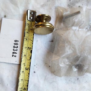 May include: A round brass cabinet knob with a stepped base, positioned next to a yellow measuring tape showing a diameter of approximately 1.25 inches. A small plastic bag containing a metal screw is included next to the knob and a white tag.