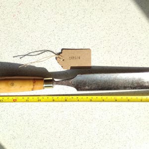 Puede incluir: Un cincel de mango de madera con una hoja de metal, que mide aproximadamente 30 cm de largo. El cincel está sobre una superficie blanca con una etiqueta de papel marrón que dice "SORL 0".