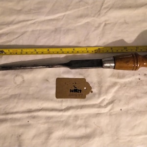 May include: A wooden handled chisel with a metal blade. The chisel is approximately 10 inches long. A yellow measuring tape is visible in the background.
