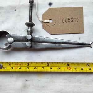 May include: Vintage metal caliper tool with a screw adjustment and a tag labeled "002559". A yellow tape measure shows inches. The caliper is approximately 6 inches long.