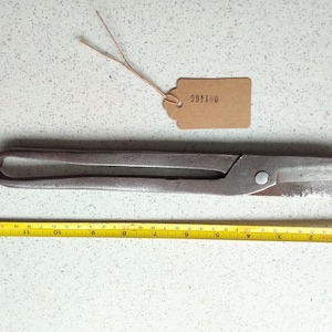 Puede incluir: Una tijera de metal vintage con una etiqueta de papel marrón que dice "999710". Las tijeras miden aproximadamente 25 cm de largo y tienen una pátina oxidada.