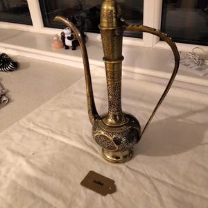 May include: A gold metal teapot with intricate floral designs. The teapot has a tall, slender body and a curved spout. The handle is attached to the top of the teapot and curves around to the side.
