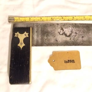 May include: A vintage metal try square with a black wooden handle and a brass inlay. The try square has a brass tag attached to it with the number "01661" written on it.