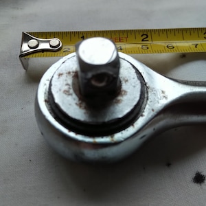 Puede incluir: Primer plano de una llave de carraca cromada con una cinta métrica amarilla. La llave mide aproximadamente 5 cm de ancho. La cinta métrica muestra medidas en pulgadas.