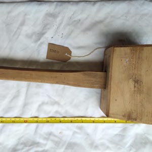 May include: A wooden mallet with a rectangular head and a long handle. The mallet is light brown and appears to be handmade. A measuring tape shows the mallet is approximately 12 inches long. A small tag is attached.