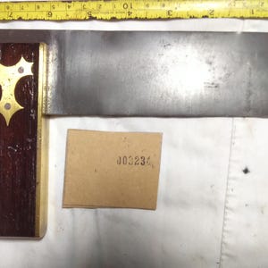 Könnte beinhalten: Ein Anschlagwinkel mit einem dunklen Holzgriff, einer Messingeinlage und einer Stahlklinge. Ein gelbes Maßband liegt über dem Werkzeug und zeigt eine Länge von etwa 23 cm. Darunter befindet sich eine kleine beige Karte mit der Nummer 003234.
