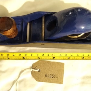 Puede incluir: Un cepillo de mano azul vintage con mango de madera. El cepillo mide aproximadamente 23 cm de largo. Una cinta métrica amarilla se coloca junto al cepillo. Una pequeña etiqueta con el número "002590" está adjunta.