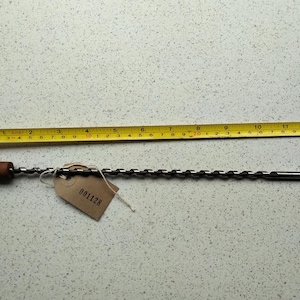 May include: A silver drill bit with a brown wooden handle, measuring approximately 12 inches in length. The drill bit is lying on a white surface with a yellow measuring tape next to it. The drill bit has a label with the number "001128" on it.