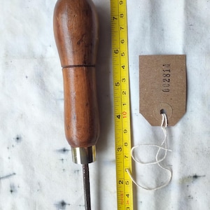 May include: A leatherworking tool with a wooden handle and a curved metal blade. The tool is approximately 15 cm long. A yellow measuring tape is next to the tool. A small brown tag with the number "002814" is attached.