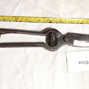 May include: A pair of metal pruning shears with a curved blade and a spring mechanism, positioned next to a yellow measuring tape showing a length of approximately 9 inches, with a small white tag labeled 003514.