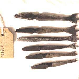 May include: A collection of seven antique metal tools, possibly for woodworking or leathercraft, arranged on a white surface. Each tool has a flat, elongated body with a pointed end and a forked or notched design at the opposite end. A tag with the number "GC2740" is attached.