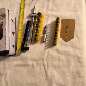 May include: A vintage tin box with a grouse illustration and the text "The Grouse" and "Perth Scotch Whisky" on a white background. The box is next to a set of yellow and black screwdrivers and a yellow and black socket wrench set. A yellow tag with the text "901606" is also in the image.