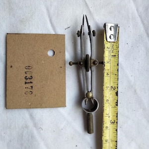 May include: A vintage metal drafting compass with a central adjustment wheel, positioned next to a yellow measuring tape showing a length of approximately 3.5 inches, alongside a rectangular tan cardboard tag printed with the number 003178.